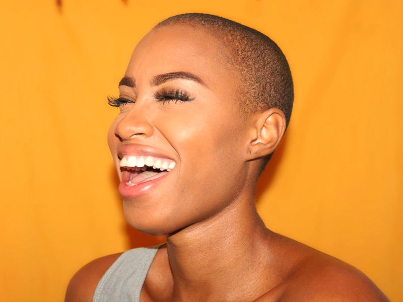young woman with shaved head laughs in front of a bright yellow background